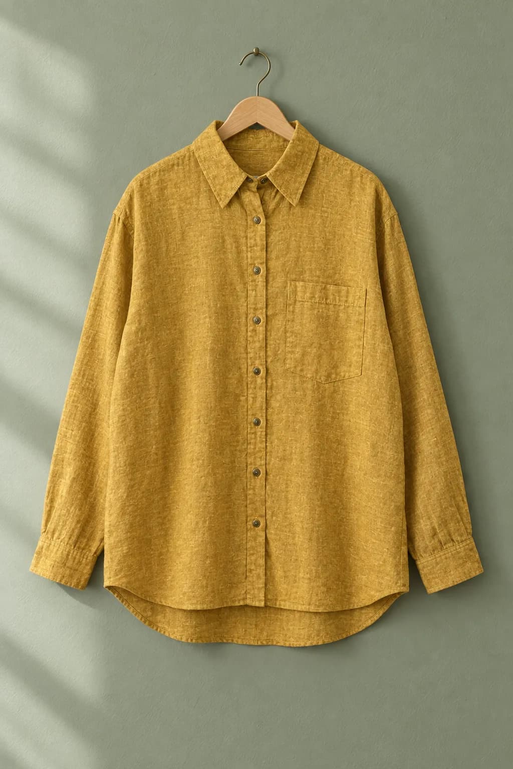 Kemeja linen mustard tergantung di depan dinding hijau sage
